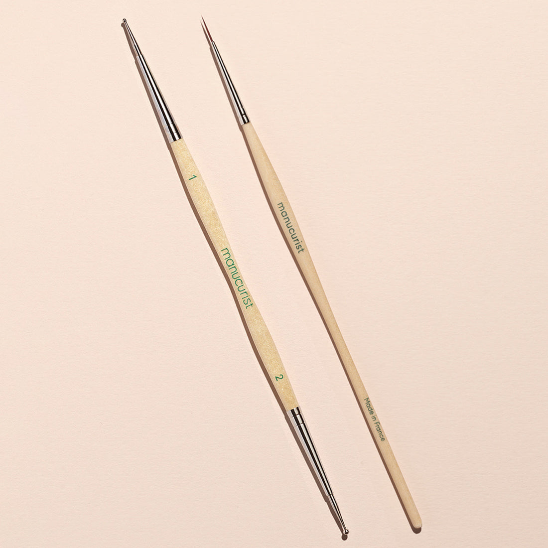 The Manucurist Dotting Tool Nail Art Liner Brush Duo features two slim, double-ended brushes with beige handles and metal ferrules, fine bristles, numbered labels, and "MANUCURIST" in green on a light pastel background.