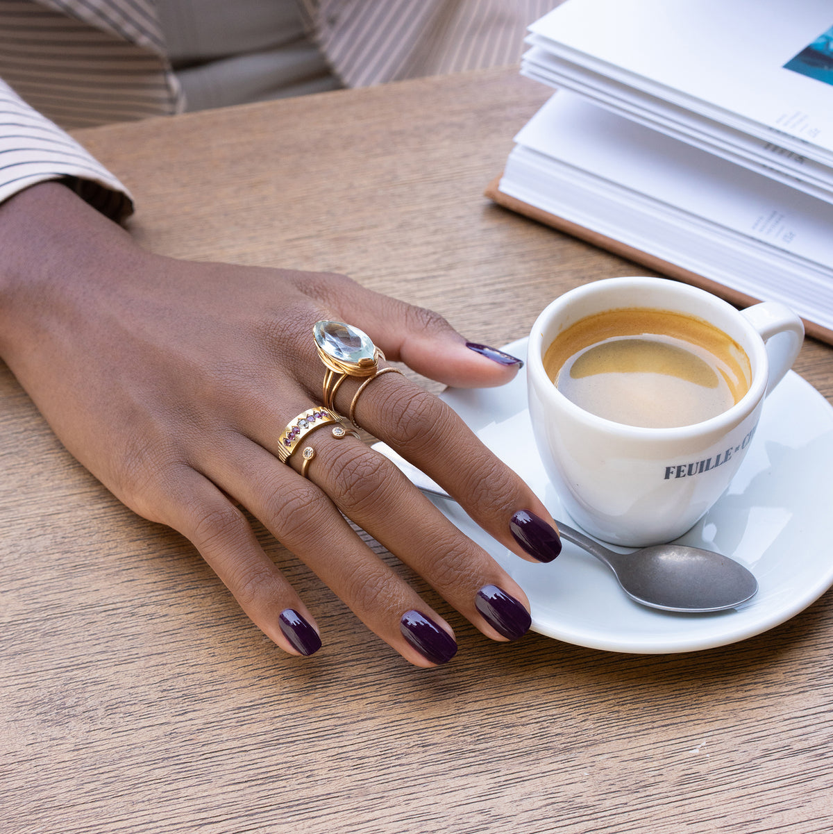 A hand with Manucurist’s Aubergine nail polish and several gold rings rests on a wooden table beside a cup of espresso, a spoon, and an open book.