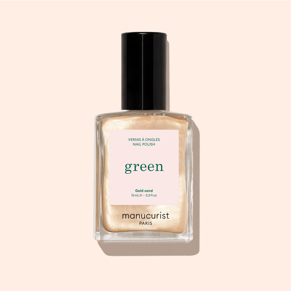 A bottle of Manucurist Gold Sand nail polish, featuring a black cap and filled with shimmering yellow lacquer, rests on a beige background.