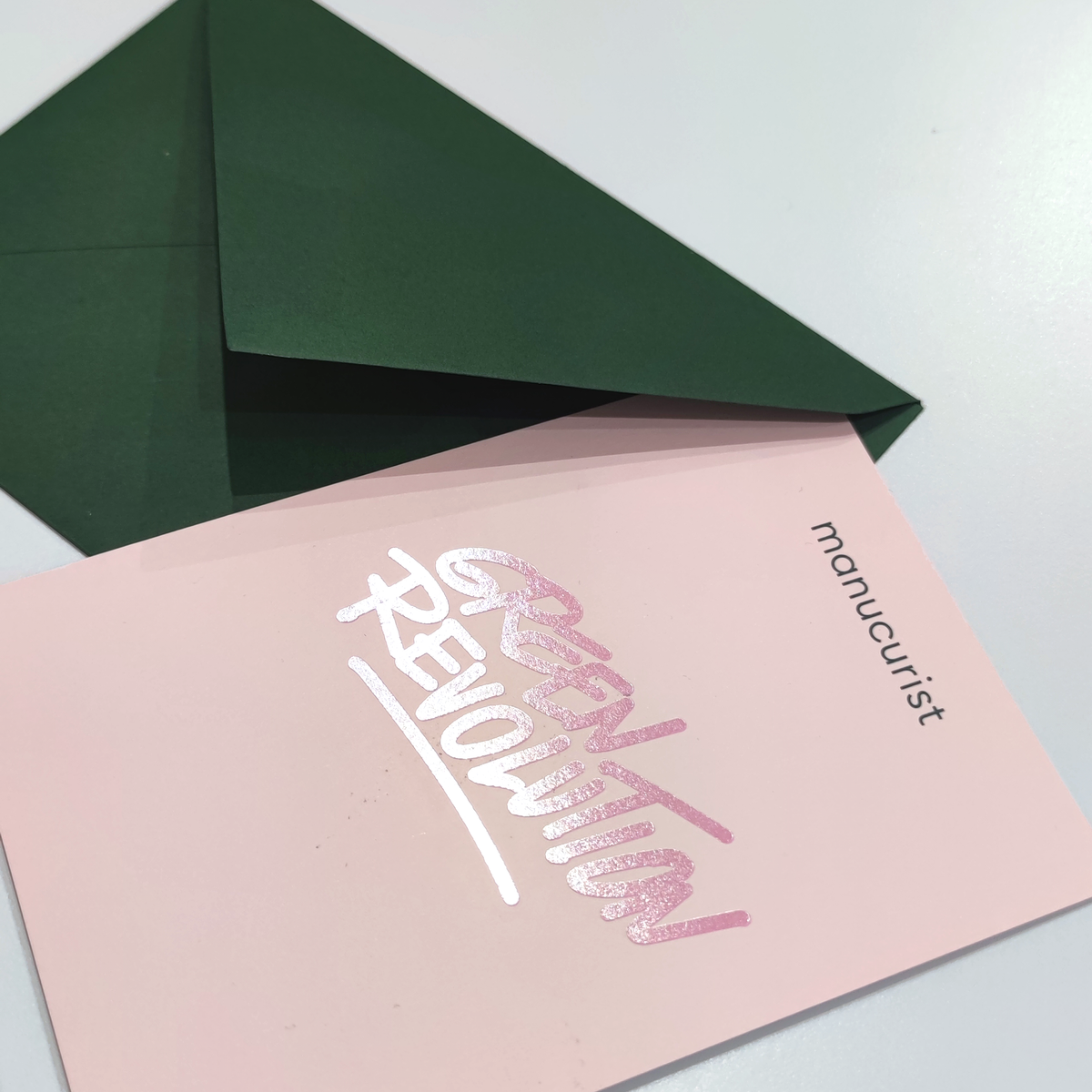 A green envelope sits beside a Manucurist Gift Card in pink with "GREEN REVOLUTION" in shiny letters—perfect for green gifting. Both rest on a white surface, with "manucurist" printed in the card’s corner.