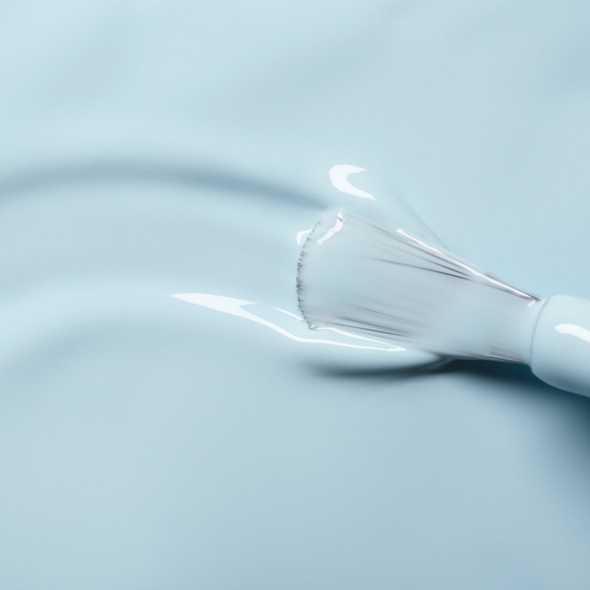 A close-up of a Manucurist Light Blue nail polish brush spreading shiny pastel blue gel on a smooth surface, with visible bristles and wet polish.