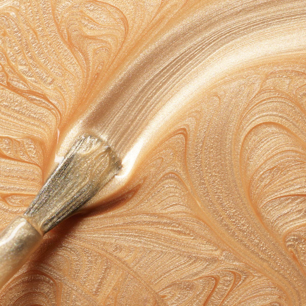 A close-up of a brush spreading Manucurist’s Gold Sand nail polish in swirling patterns on a surface, creating the luminous effect of shimmery, metallic gold.