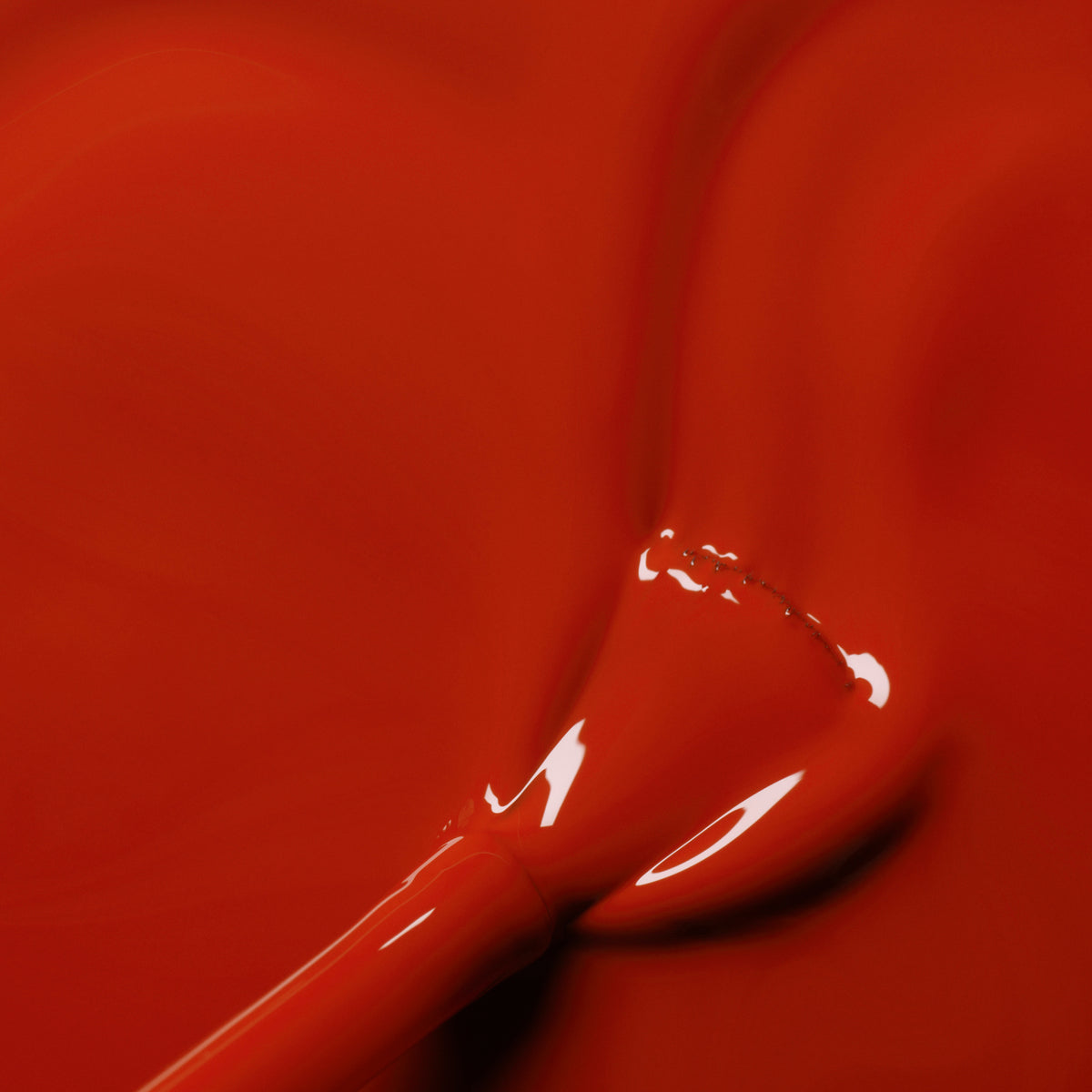 A close-up of a Manucurist Indian Summer nail polish brush coated with shiny brick red polish, resting on a smooth surface with the same rich red shade spread across it.