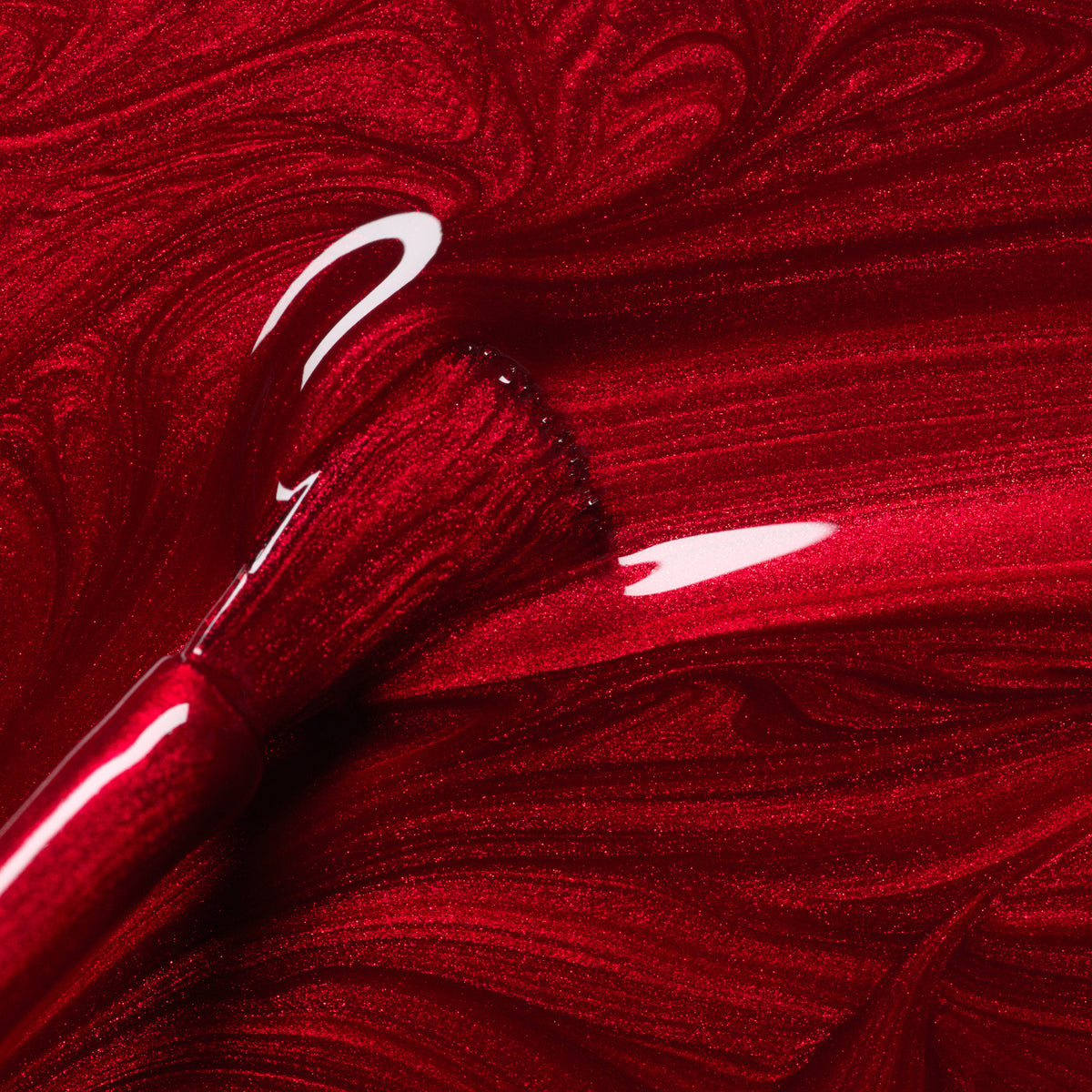A close-up of a brush coated in Manucurist’s Red Hibiscus gel polish, spreading the glossy, iridescent red shade in swirling patterns across a smooth surface.