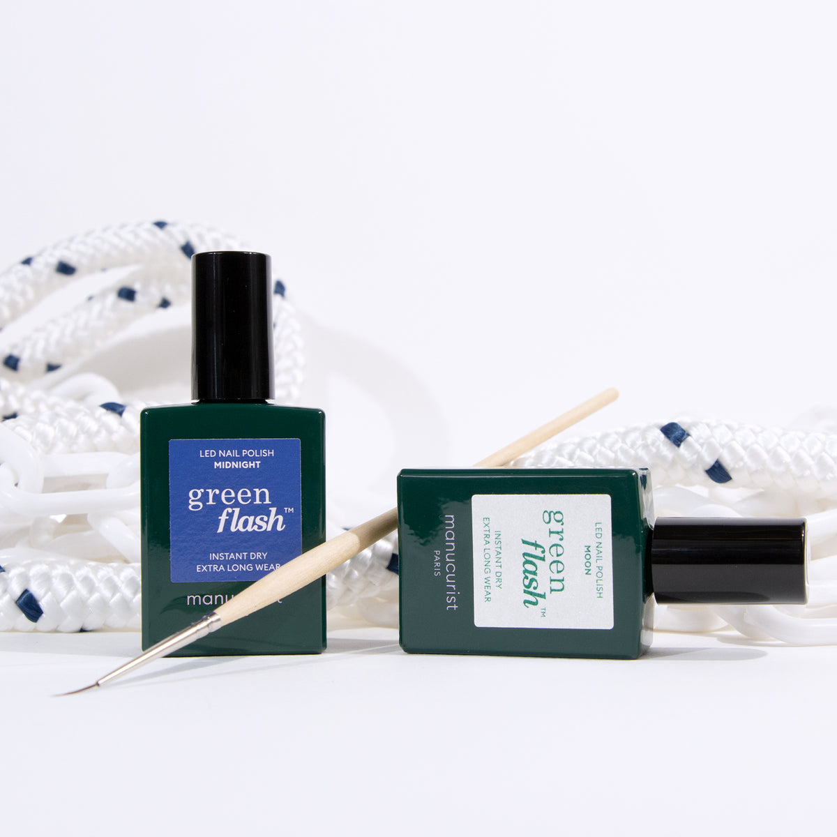 Two bottles of Manucurist Midnight Swim Green Flash LED nail polish—one upright, one lying down—are paired with a wooden cuticle stick and a coiled white rope with blue accents, ideal for nautical-inspired nail art.