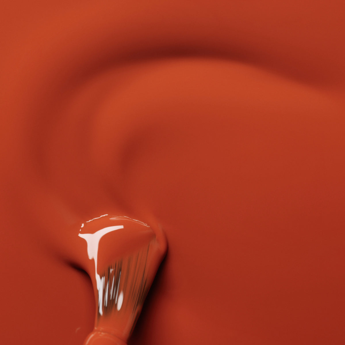 A close-up of a paintbrush spreading Manucurist Terracotta gel polish in swirling strokes, highlighting the glossy finish and reflecting the warmth of scorched red earth. Bristles and polish shine vividly in the shot.