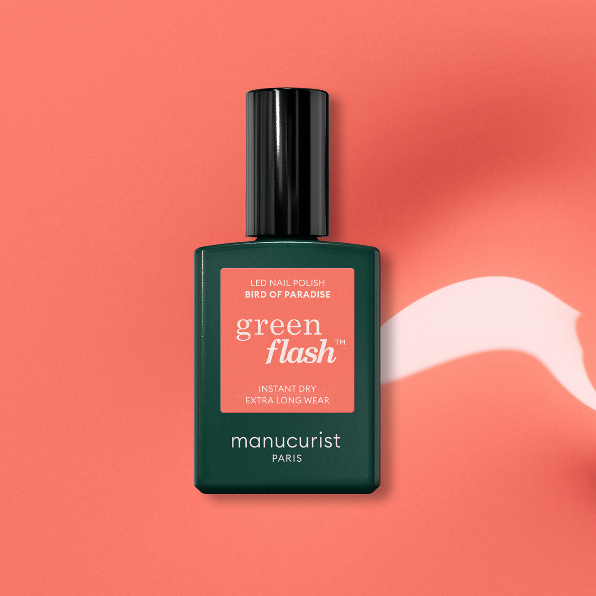 A bottle of Manucurist Bird of Paradise gel polish rests on a coral pink background with a white paint streak. The dark green bottle features a coral label, reflecting the vibrant quality of this salon-worthy shade.