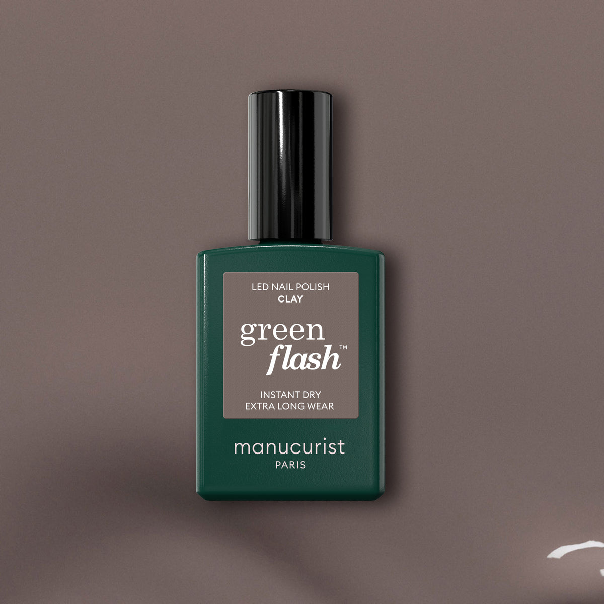 A bottle of Manucurist Green Flash gel polish in the shade Clay stands upright on a brown background, featuring a dark green bottle with a rectangular label—ideal for lovers of taupe gel polish.