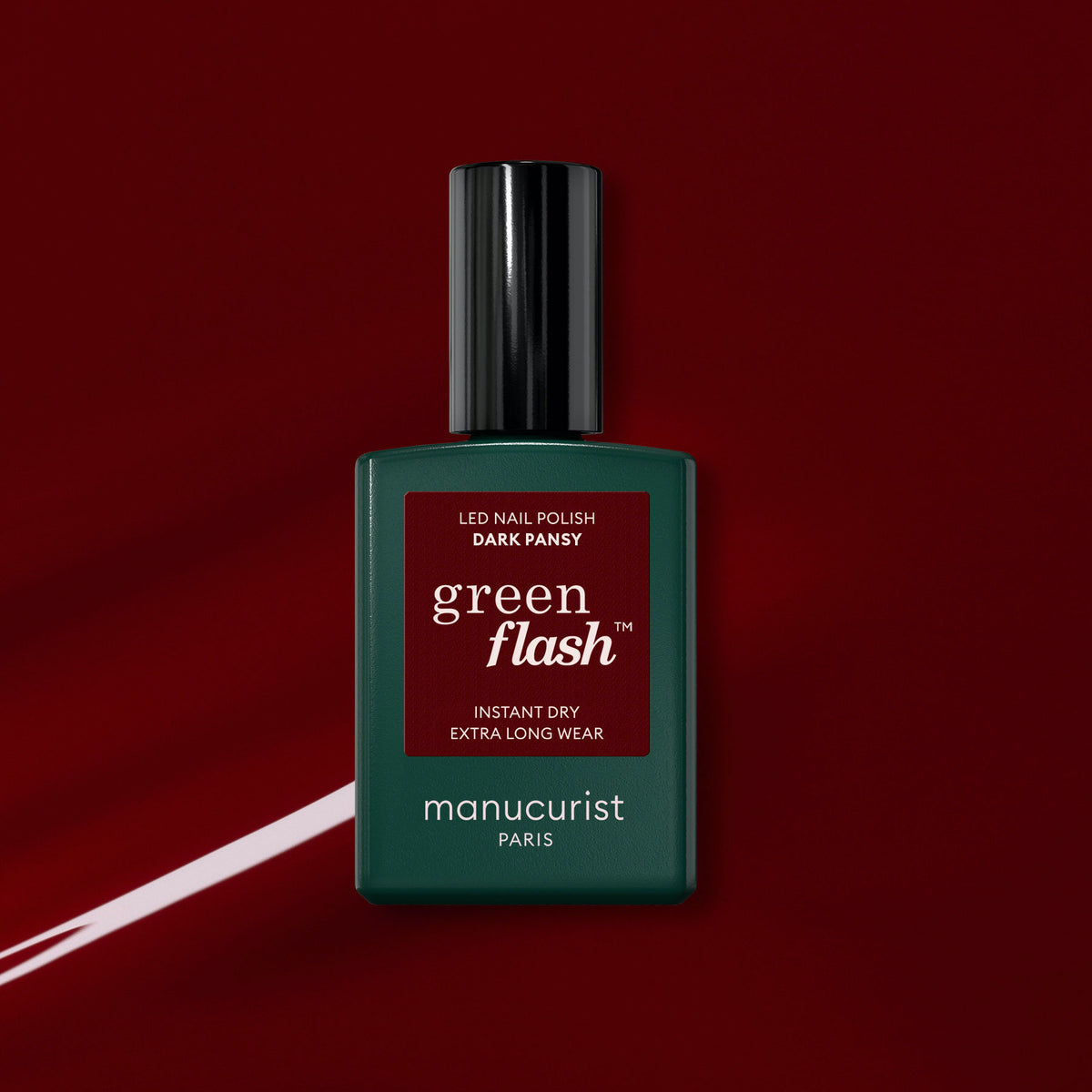 A bottle of Manucurist Dark Pansy Green Flash gel polish is displayed against a glossy red nail polish background, featuring a dark green square bottle with a black cap.