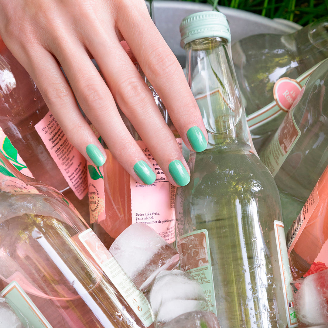 A hand with Amande by Manucurist almond green nail polish rests on glass bottles of pink and clear drinks in an ice-filled metal tub.