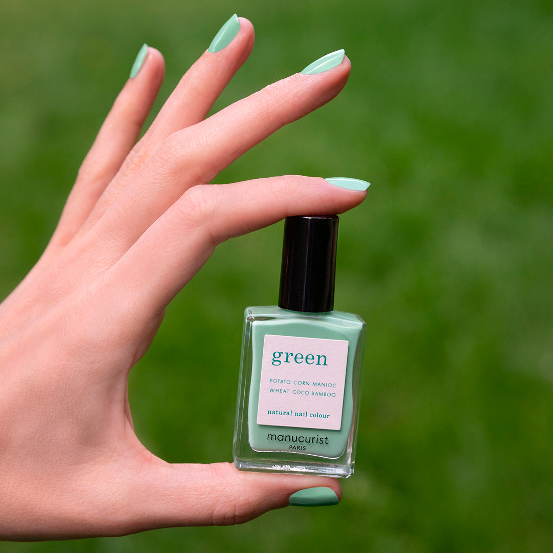 A hand with neatly manicured almond green nails holds a Manucurist Amande nail polish bottle. The background is a blurred grassy area.