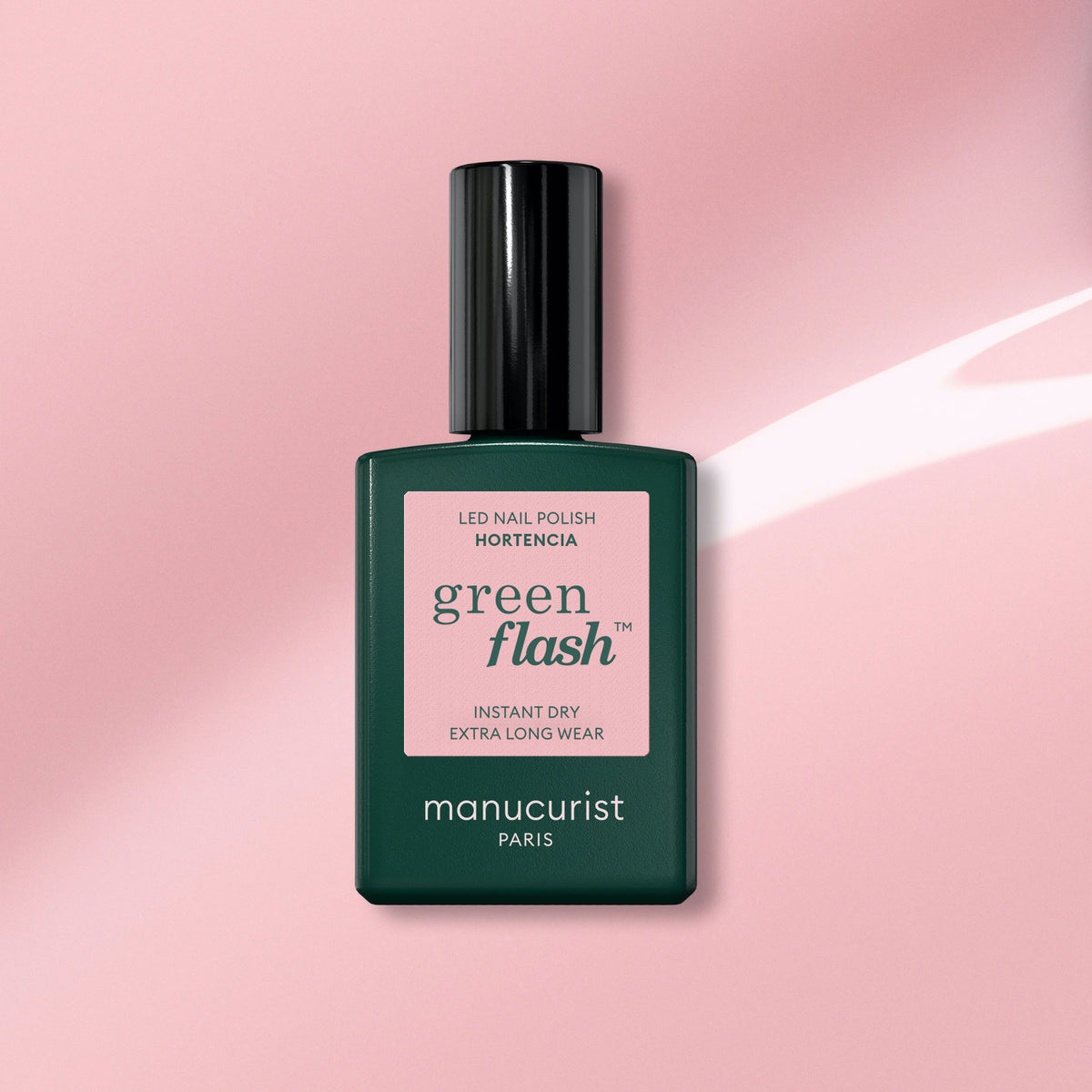A bottle of Manucurist’s Green Flash gel polish in the shade Hortencia appears on a glossy pink background. The dark green bottle displays a pink label, emphasizing this natural-hued nail color.