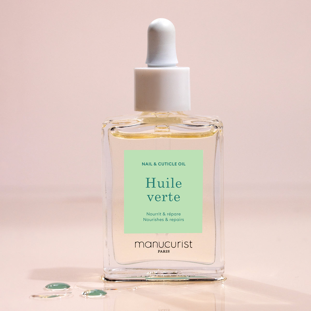 A clear glass dropper bottle of Manucurist Nourishing Cuticle Oil stands upright on a pink surface, with drops of hydrating oil nearby for nourishing nails.