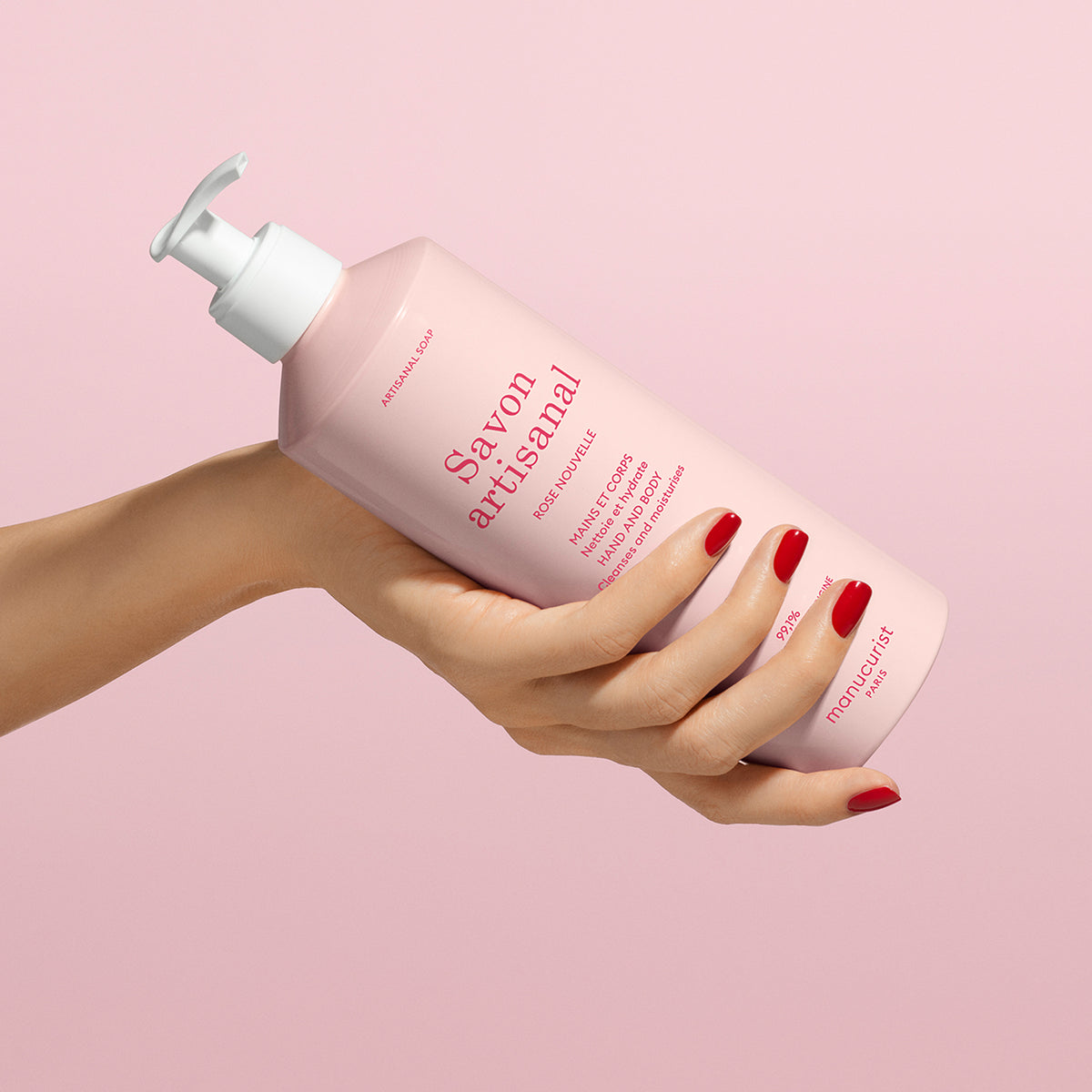 A hand with red polished nails holds a pink bottle of Manucurist Rose Nouvelle Artisan Soap - Cosmos Organic (500ml) with a pump dispenser against a matching pink background.