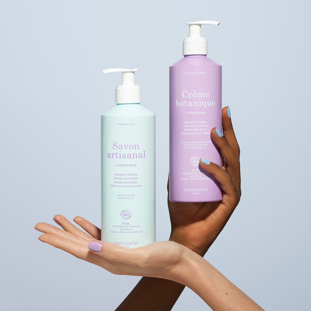 Two hands hold pastel pump bottles—one mint green labeled Manucurist and one lavender labeled Soothing Lavande Hand  Body Cream - Cosmos Organic—against a light blue background. Both are for hand and body care.