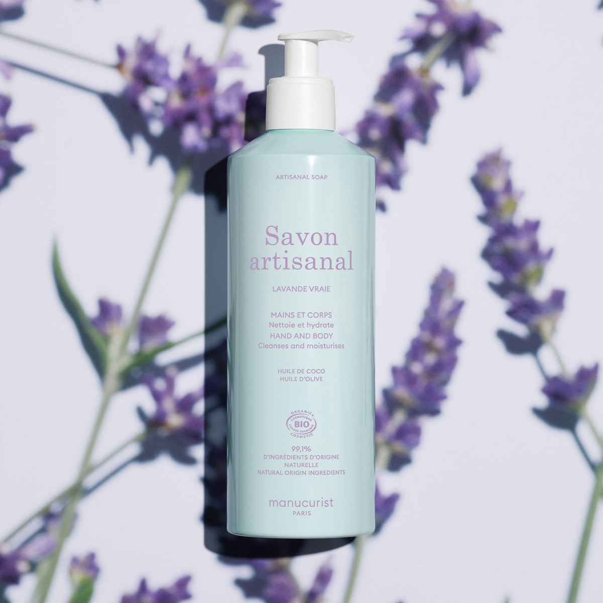 A pale blue 500ml bottle of Manucurist Lavande Artisan Soap - Cosmos Organic with a pump sits among lavender flowers, its label emphasizing organic and natural ingredients.