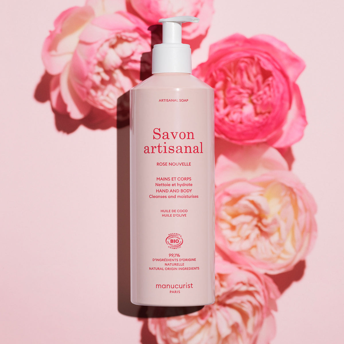 A pink bottle of Manucurist Rose Nouvelle Artisan Soap (Cosmos Organic, 500ml) sits on a pink surface with roses. The label highlights natural ingredients, moisturizing benefits, and a delicate rose fragrance.