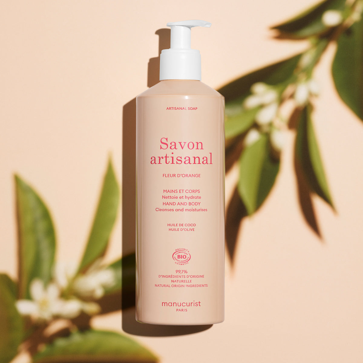 A beige pump bottle of Manucurist “Fleur d’Orange” Artisan Soap (Cosmos Organic, 500ml) with red text sits on a pale surface, backed by green leaves and shadows. The label highlights natural ingredients and Orange Blossom scent.
