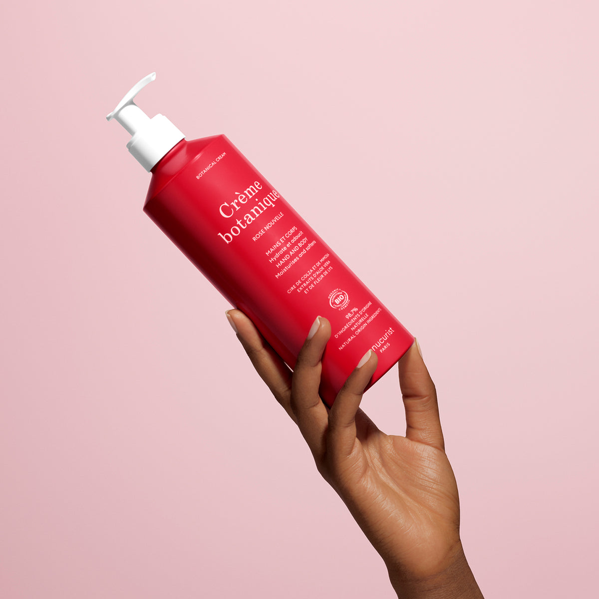 A hand holds a red 500ml bottle labeled "Rose Nouvelle Cream - Cosmos Organic" by Manucurist with white text and a white pump, set against a light pink background. The product appears to be organic skincare or hand cream.