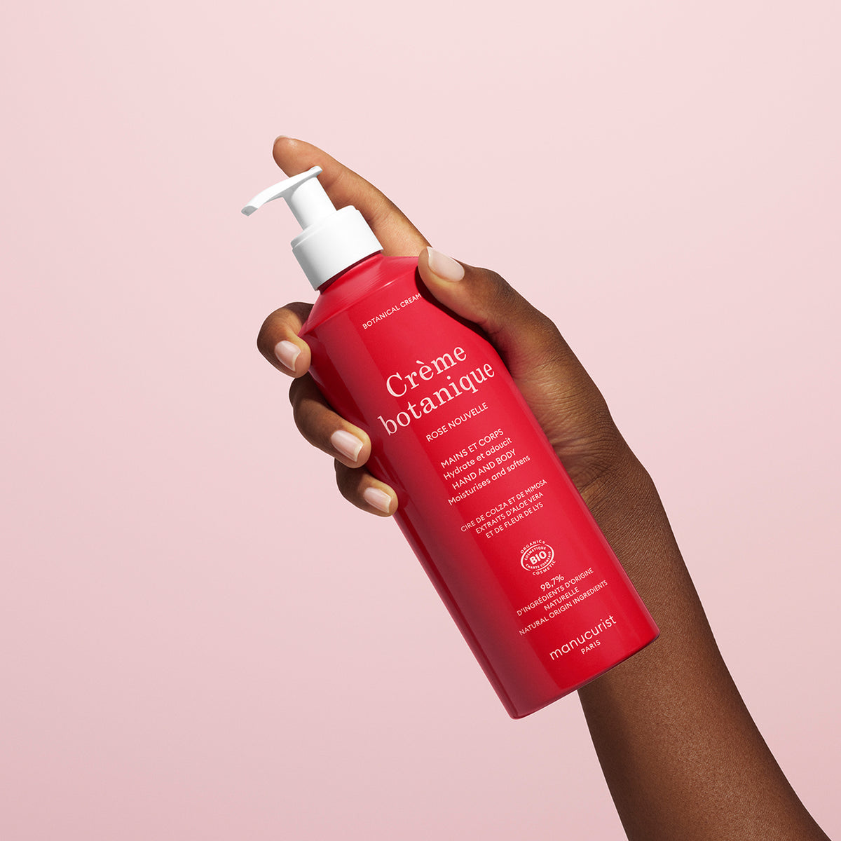 A hand holds a red bottle of Manucurist Rose Nouvelle Cream - Cosmos Organic (500ml) with a white pump, against a light pink background. The label shows white text and botanical-themed graphics.