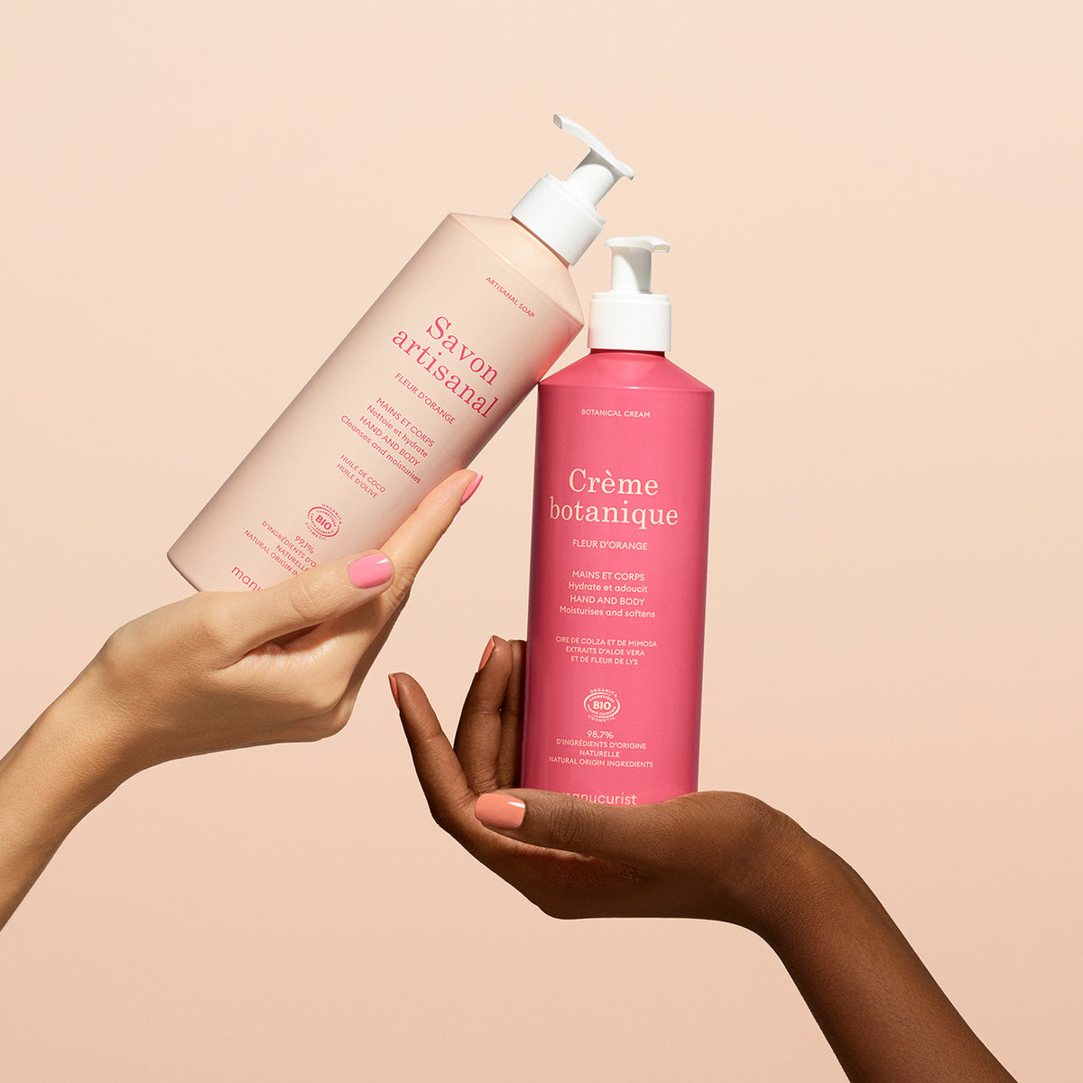 Two hands hold up pink bottles with pump dispensers against a beige background; one light pink bottle labeled Manucurist, the other dark pink, Soothing Fleur d&