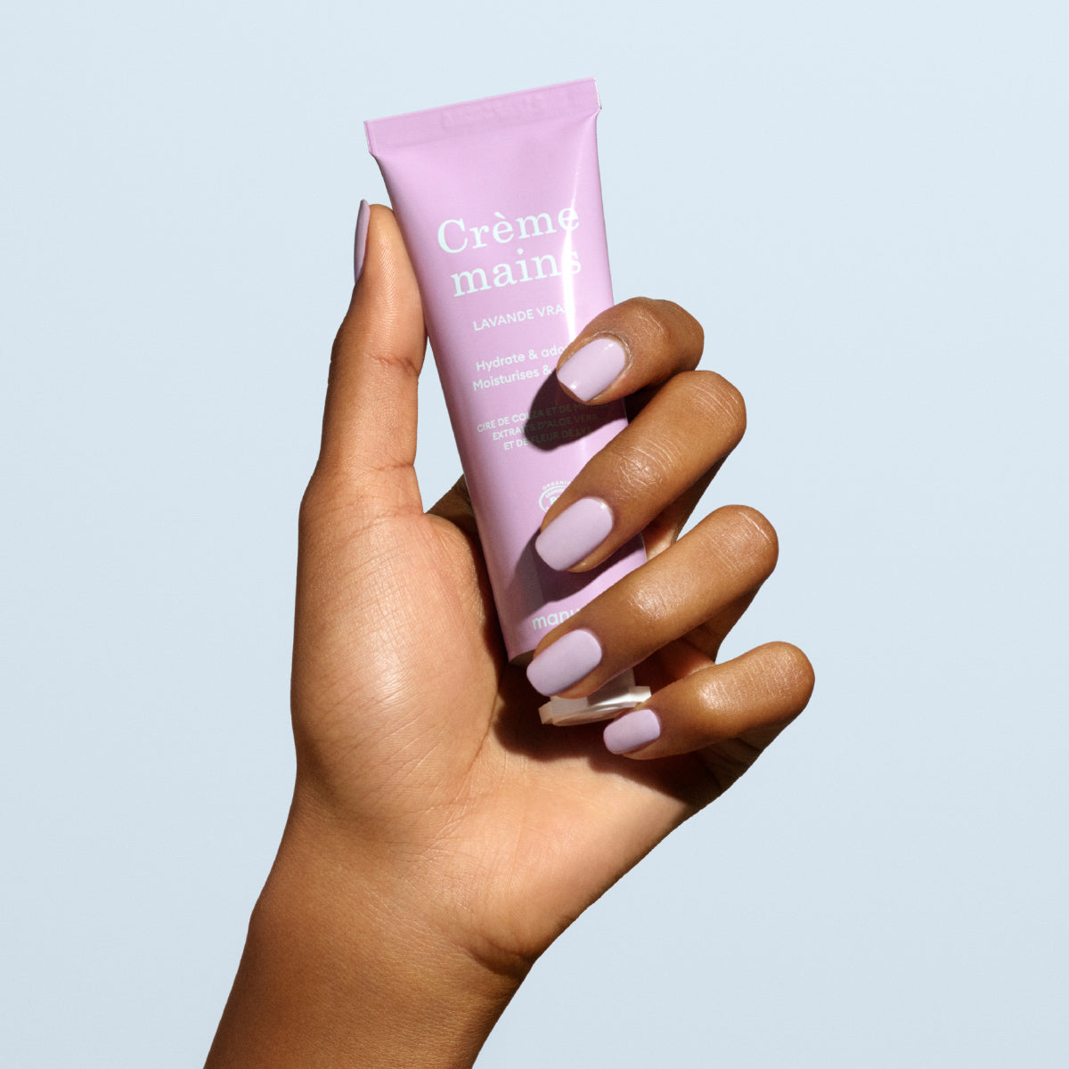 A hand with light pink nails holds a lavender tube labeled "Lavande Vraie Hand Cream - Cosmos Organic" by Manucurist, highlighting this soothing, moisturising hand cream against a pale blue background.