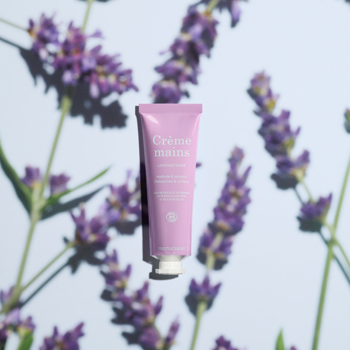 A pink tube of Manucurist Lavande Vraie Hand Cream - Cosmos Organic is centered on a background patterned with purple lavender flowers and green stems, casting soft shadows.
