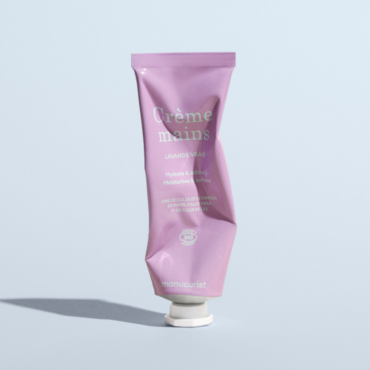 A crumpled pink tube of Manucurist Lavande Vraie Hand Cream - Cosmos Organic stands upright on a light surface, featuring white and green text on the packaging and casting a shadow to the left.