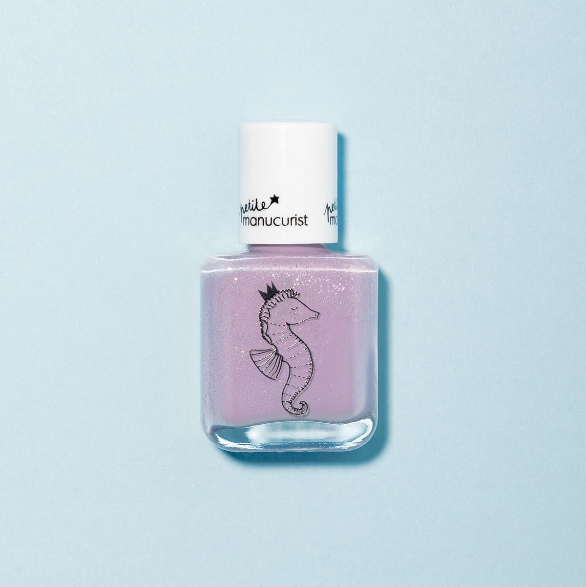 A bottle of Manucurist’s Janis the Seahorse kids’ nail polish—light purple with a white cap and a crowned seahorse illustration—sits on a pale blue background. Made from natural ingredients for safe, playful fun.