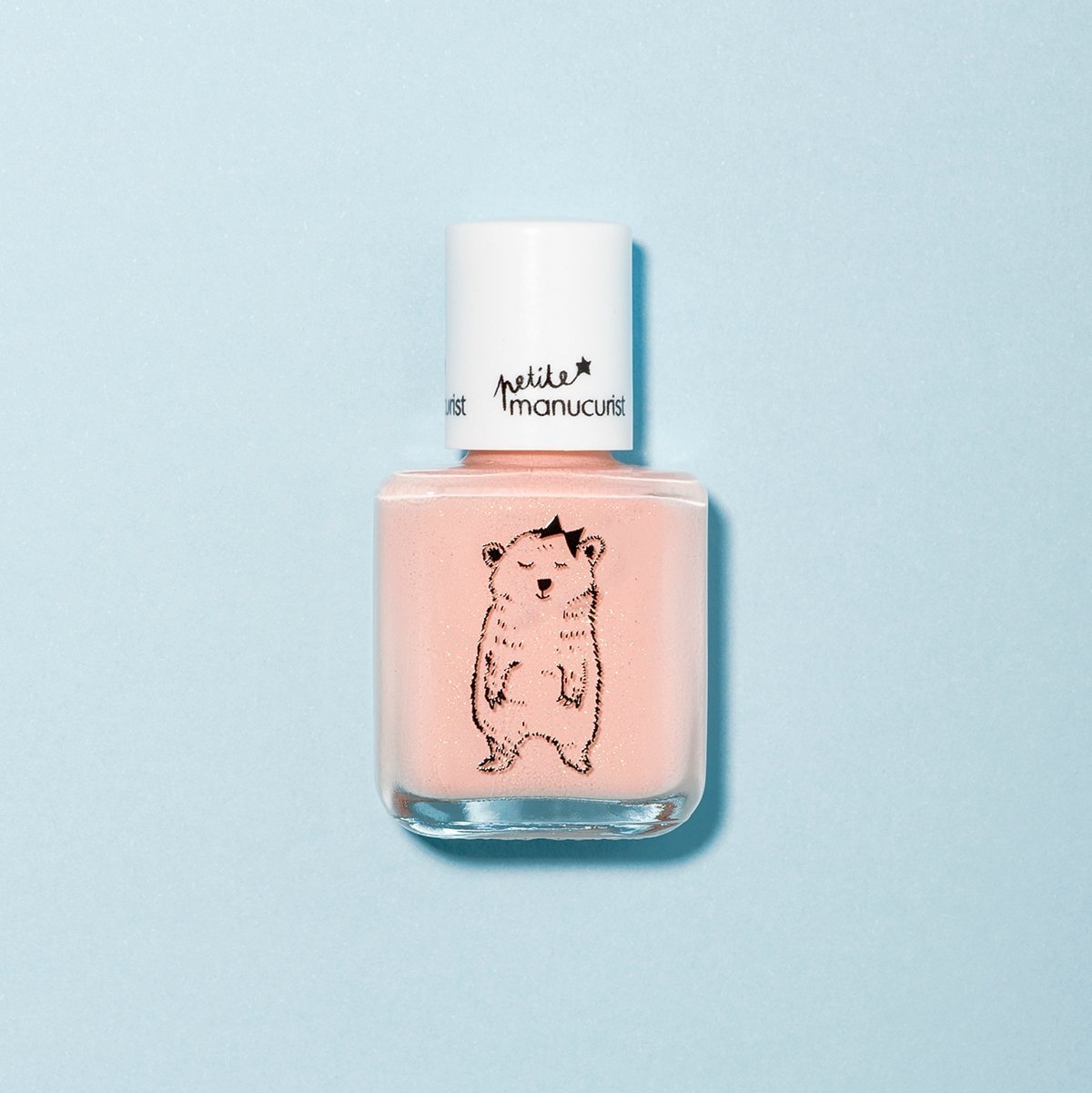 A bottle of Manucurist Joy the Bear Cub pale pink kids’ nail polish with a white cap sits on a light blue background, featuring a standing bear illustration on the front and made with naturally sourced ingredients.