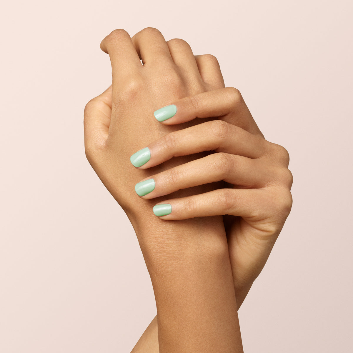 Two hands with light brown skin are crossed gracefully against a light beige background. The nails are neatly manicured and painted with Mint pastel green nail polish from Manucurist.