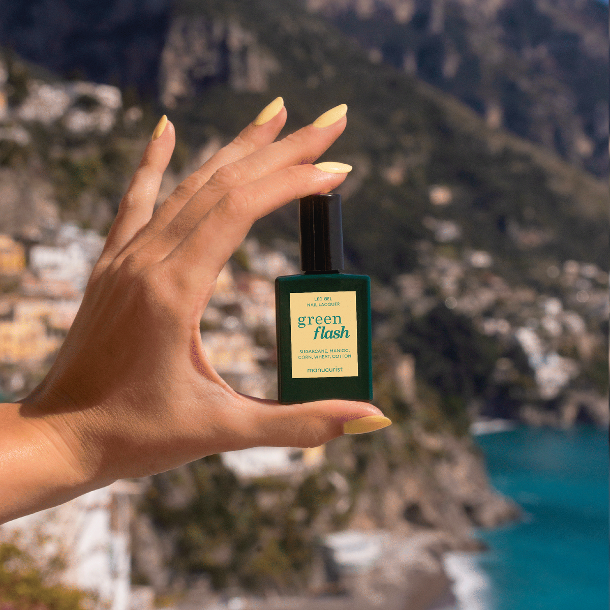A hand with pastel yellow Manucurist Mimosa gel polish holds a Green Flash bottle against a blurred coastal cliff and sea backdrop.