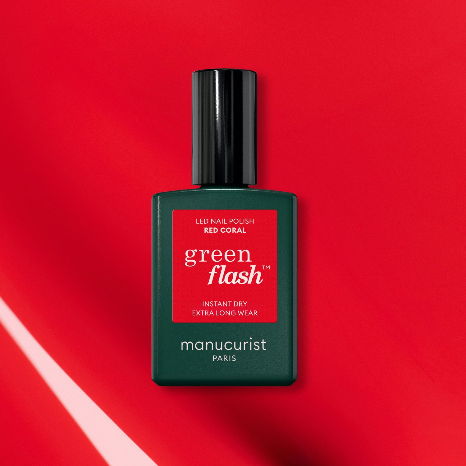 A green bottle of Manucurist-test Red Coral OLD LED nail polish is displayed on a vibrant red background with light reflections, showcasing its long-lasting gel nail polish formula.