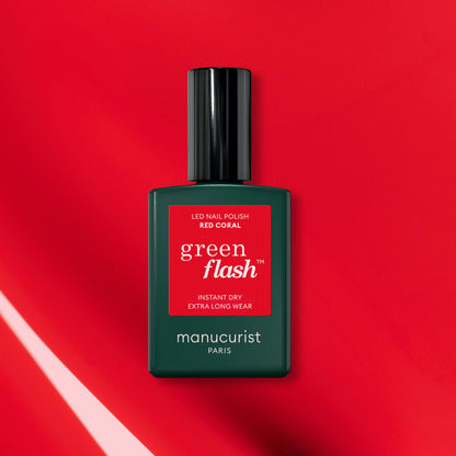 A green bottle of Manucurist-test Red Coral OLD LED nail polish is displayed on a vibrant red background with light reflections, showcasing its long-lasting gel nail polish formula.