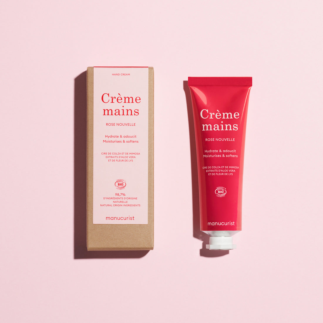 A pink tube and cardboard box of Manucurist Rose Nouvelle Hand Cream - Cosmos Organic, with a delicate rose scent and nourishing benefits, are displayed on a light pink background.