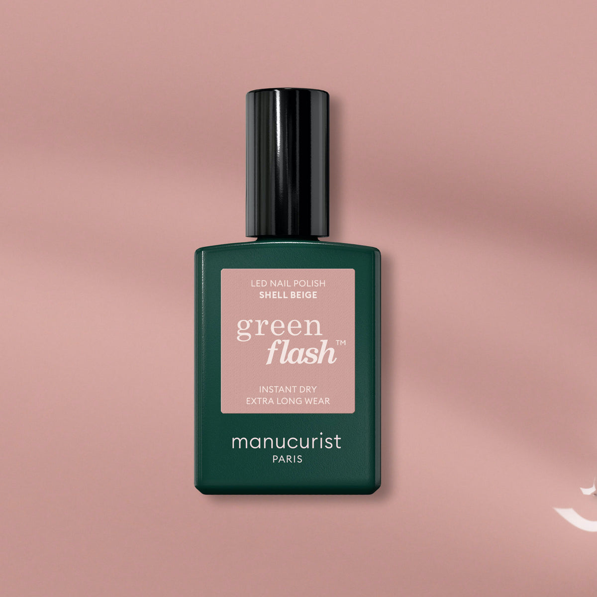 A bottle of Manucurist Shell Beige gel polish with a dark green cap and label rests on a blush pink background, highlighting its delicate pinkish beige shade.