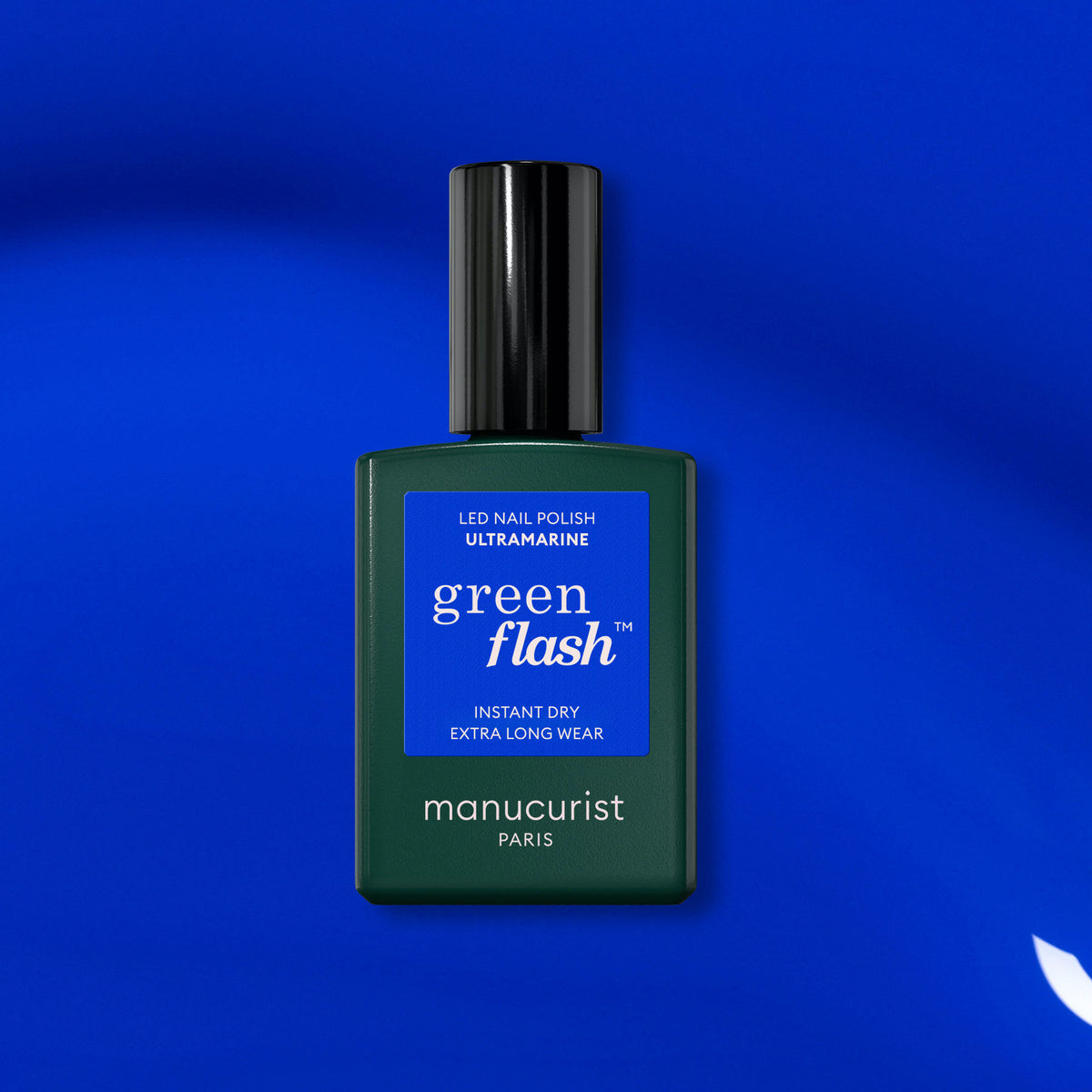 A bottle of Manucurist Ultramarine gel polish sits on a vibrant blue backdrop. The dark green bottle with a black cap and blue label pairs perfectly with the rich blue shade inside.