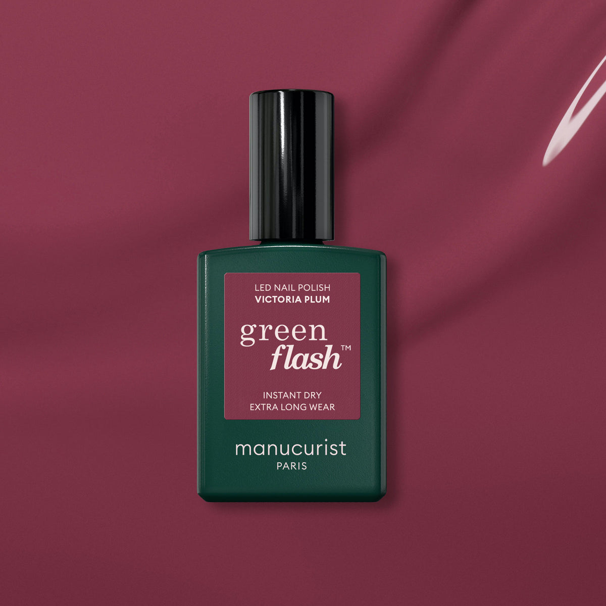 A bottle of Manucurist Victoria Plum gel polish is displayed on a plum-colored background, featuring the brand’s signature dark green bottle with a black cap and square label.