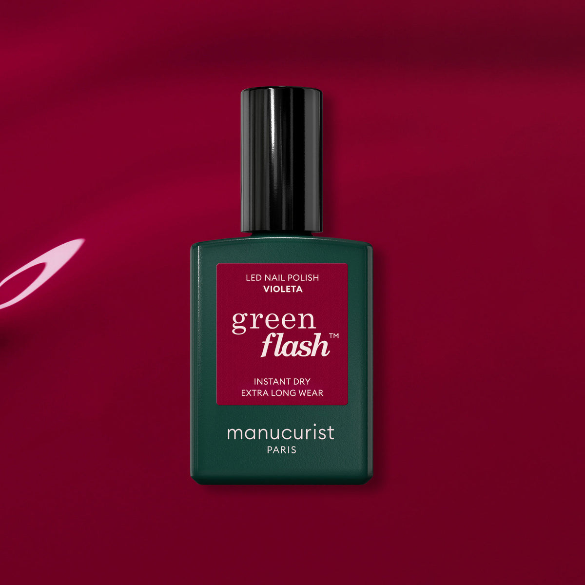 A bottle of Manucurist Violeta sits on a glossy, deep magenta surface. The dark green bottle with a black cap and magenta label displays this vibrant pinky purple gel polish.